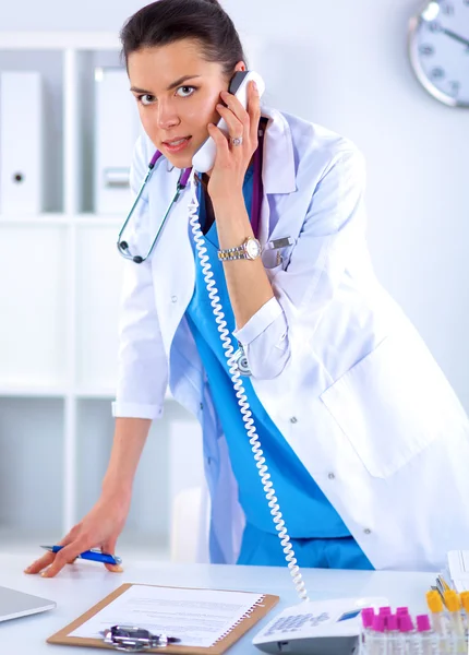 Young woman doctor in white coat at computer using phone — Stock Photo, Image