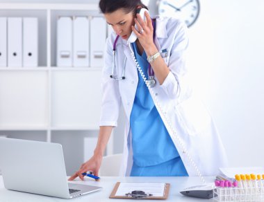 Bilgisayarda telefon kullanan beyaz önlüklü genç kadın doktor.