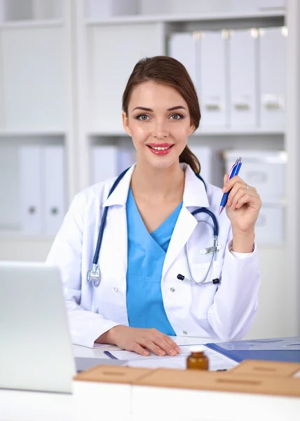 Hermosa joven sonriente doctora sentada en el escritorio y escribiendo. —  Fotos de Stock