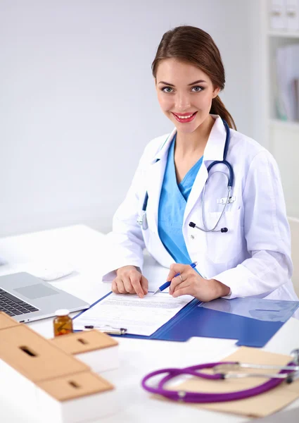 Bonito jovem sorridente médico feminino sentado na mesa e escrevendo. — Fotografia de Stock