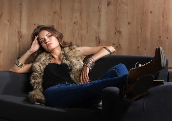 Porträtt av elegant kvinna som sitter på svart soffa klädd i en blå jeans och päls väst — Stockfoto