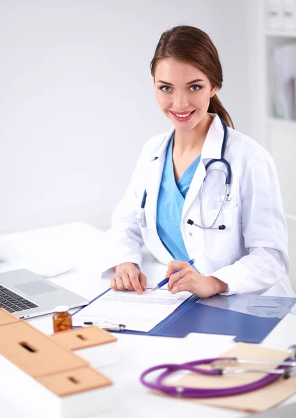 Hermosa joven sonriente doctora sentada en el escritorio y escribiendo. — Foto de Stock