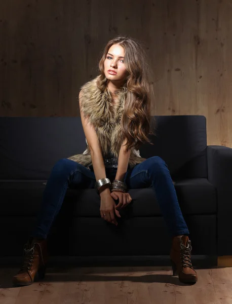 Retrato de mulher elegante sentado no sofá preto vestindo uma calça azul e colete de pele — Fotografia de Stock