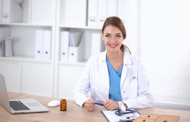 Beautiful young smiling female doctor sitting at the desk and writing. clipart