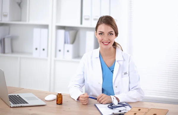 Όμορφη νεαρή χαμογελαστή γυναίκα γιατρός κάθεται στο γραφείο και γράφει. — Φωτογραφία Αρχείου