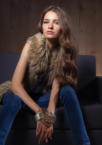 Retrato de una mujer elegante sentada en un sofá negro con pantalones vaqueros azules y chaleco de piel — Foto de Stock