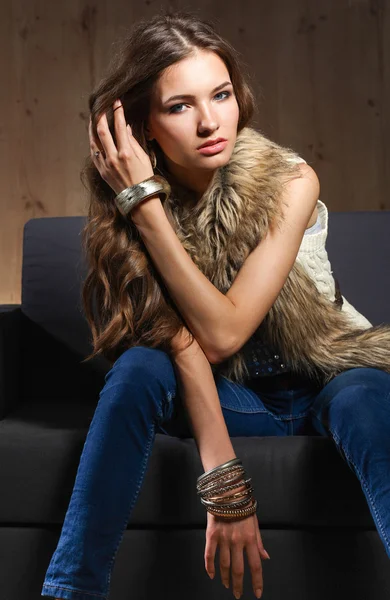 Portrait of elegant woman sitting on black sofa wearing a blue jeans and fur vest — Stock Photo, Image