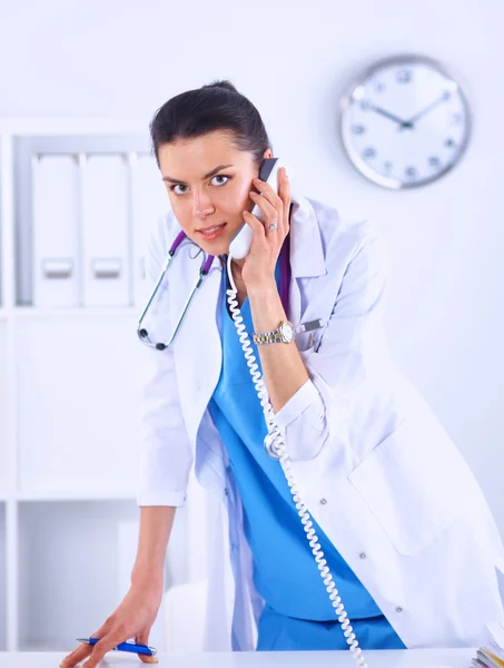 Young woman doctor in white coat at computer using phone — Stock Photo, Image