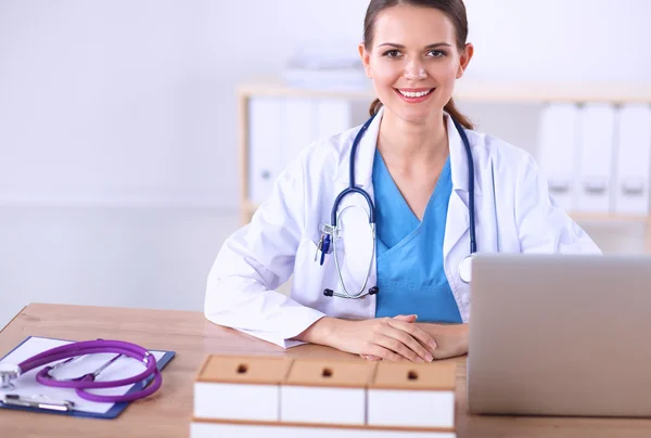 Mooie jonge glimlachende vrouwelijke arts zitten aan het bureau en schrijven. — Stockfoto