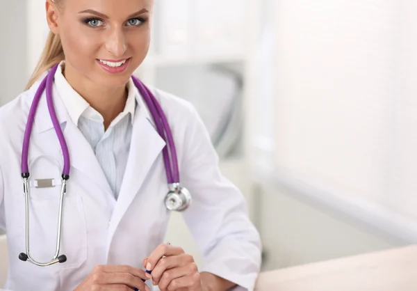 Bonito jovem sorridente médico feminino sentado na mesa e escrevendo. — Fotografia de Stock