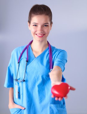 Kırmızı bir kalp taşıyan genç bir kadın doktor, gri arka planda duruyor.
