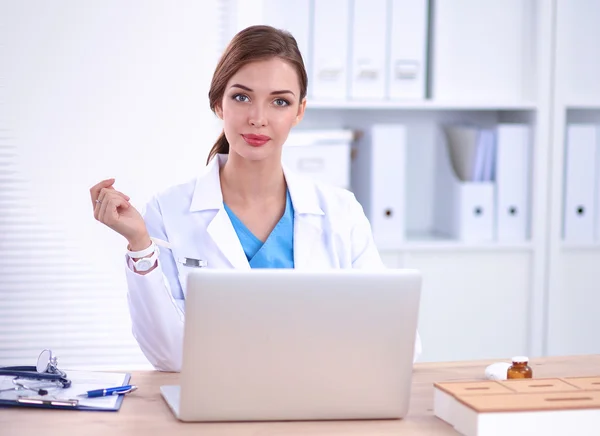 Schöne junge lächelnde Ärztin sitzt am Schreibtisch und schreibt. — Stockfoto