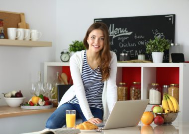 Dizüstü bilgisayar kullanarak ve mutfakta oturmuş çekici genç kadın 