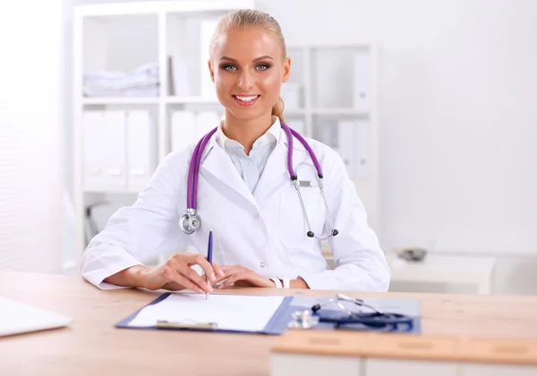 Mooie jonge glimlachende vrouwelijke arts zitten aan het bureau en schrijven. — Stockfoto