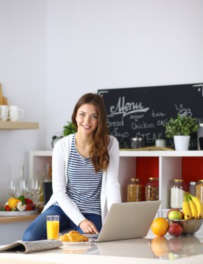 Dizüstü bilgisayar kullanarak ve mutfakta oturmuş çekici genç kadın 