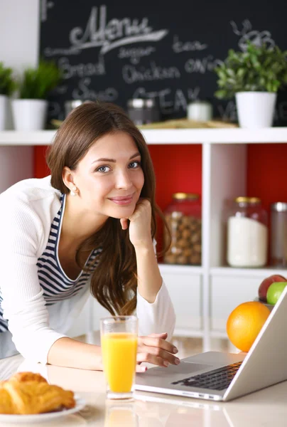 Ελκυστική νεαρή γυναίκα χρησιμοποιώντας φορητό υπολογιστή και να κάθεται στην κουζίνα — Φωτογραφία Αρχείου