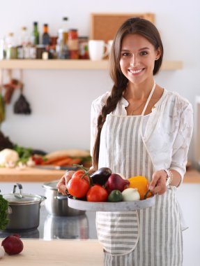 Mutfakta elinde sebzelerle gülümseyen genç bir kadın.