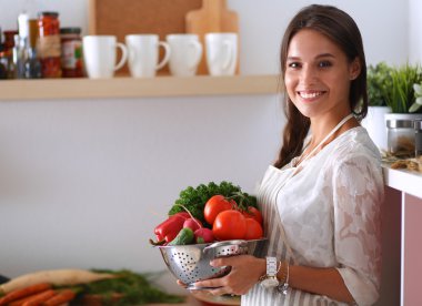 Mutfakta elinde sebzelerle gülümseyen genç bir kadın.