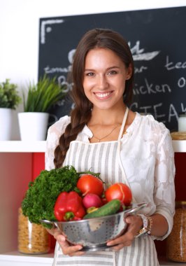 Mutfakta elinde sebzelerle gülümseyen genç bir kadın.