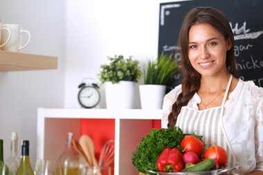 Mutfakta elinde sebzelerle gülümseyen genç bir kadın.