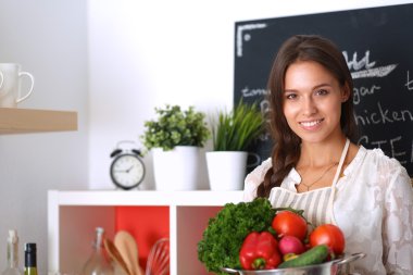 Mutfakta elinde sebzelerle gülümseyen genç bir kadın.