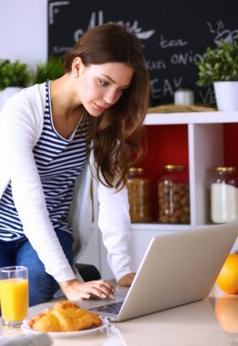 Dizüstü bilgisayar kullanarak ve mutfakta oturmuş çekici genç kadın 