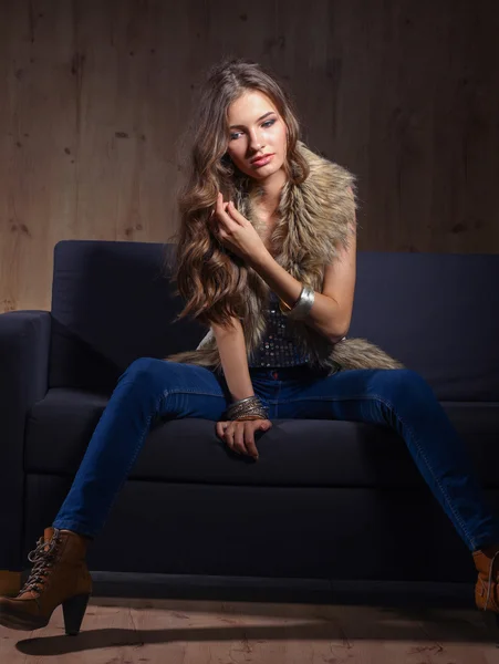 Ritratto di donna elegante seduta sul divano nero con jeans blu e gilet di pelliccia — Foto Stock