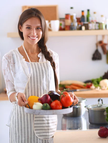 Mujer joven sonriente sosteniendo verduras de pie en la cocina —  Fotos de Stock