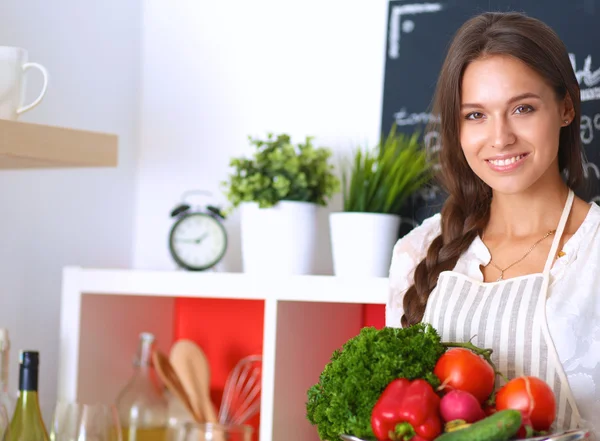 Mujer joven sonriente sosteniendo verduras de pie en la cocina —  Fotos de Stock