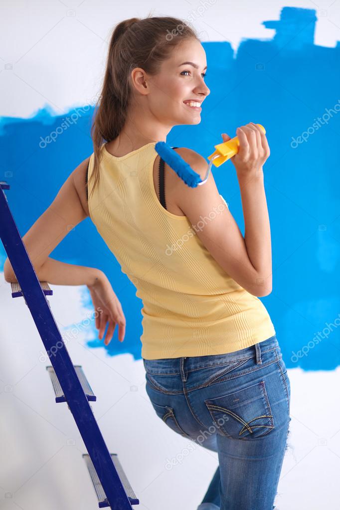Happy beautiful young woman doing wall painting, standing near ladder