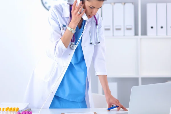 Young woman doctor in white coat at computer using phone — Stock Photo, Image