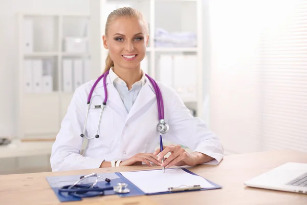 Beautiful young smiling female doctor sitting at the desk and writing. — Stock Photo, Image