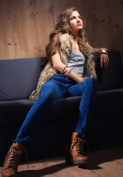 Porträt einer eleganten Frau, die auf einem schwarzen Sofa sitzt und eine blaue Jeans und Pelzweste trägt — Stockfoto