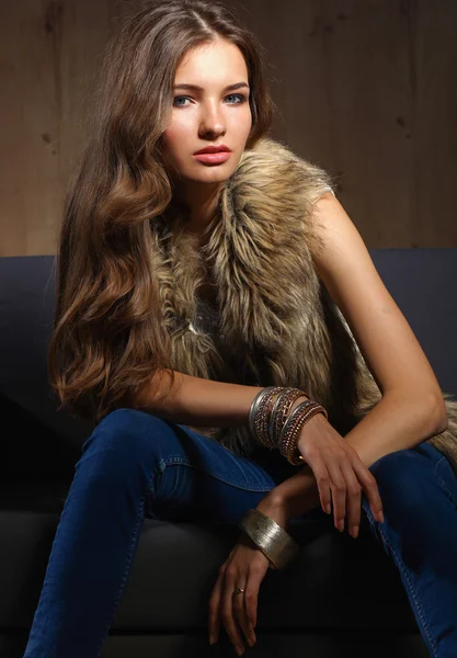 Retrato de una mujer elegante sentada en un sofá negro con pantalones vaqueros azules y chaleco de piel — Foto de Stock