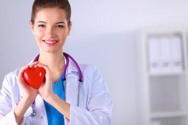 Kırmızı bir kalp taşıyan genç bir kadın doktor, gri arka planda duruyor.
