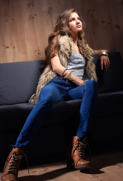 Porträt einer eleganten Frau, die auf einem schwarzen Sofa sitzt und eine blaue Jeans und Pelzweste trägt — Stockfoto