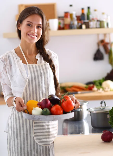 Mujer joven sonriente sosteniendo verduras de pie en la cocina —  Fotos de Stock