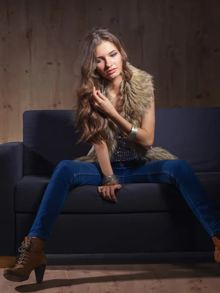 Portrait of elegant woman sitting on black sofa wearing a blue jeans and fur vest — Stock Photo, Image