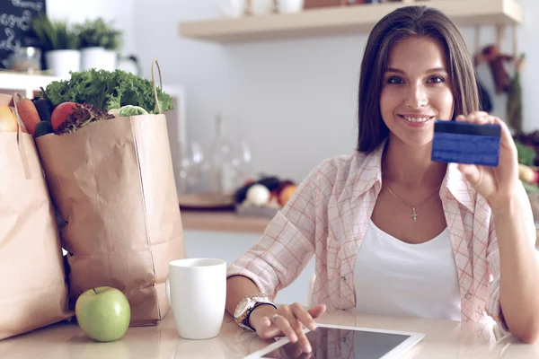 Mujer sonriente compras en línea utilizando la tableta y la tarjeta de crédito en la cocina — Foto de Stock