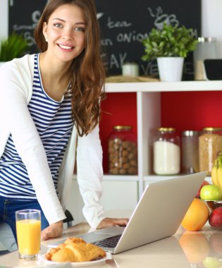 Dizüstü bilgisayar kullanarak ve mutfakta oturmuş çekici genç kadın