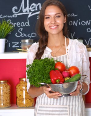 Mutfakta elinde sebzelerle gülümseyen genç bir kadın.