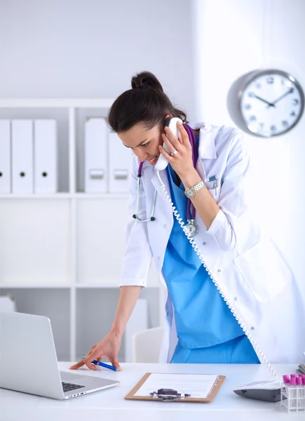 Mujer joven médico en bata blanca en el ordenador usando el teléfono —  Fotos de Stock