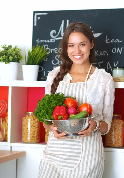 Mujer joven sonriente sosteniendo verduras de pie en la cocina —  Fotos de Stock