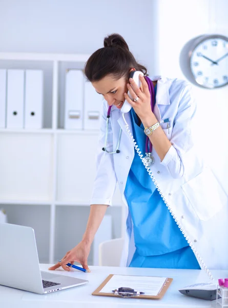 Young woman doctor in white coat at computer using phone — Stock Photo, Image