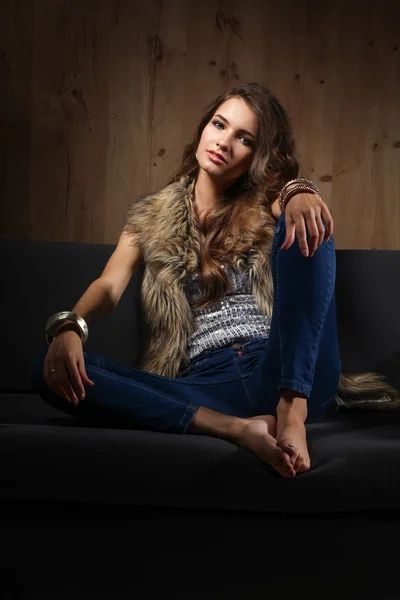 Portret elegancki kobieta siedzi na kanapie czarne noszenia jeansów i kamizelka futro — Zdjęcie stockowe
