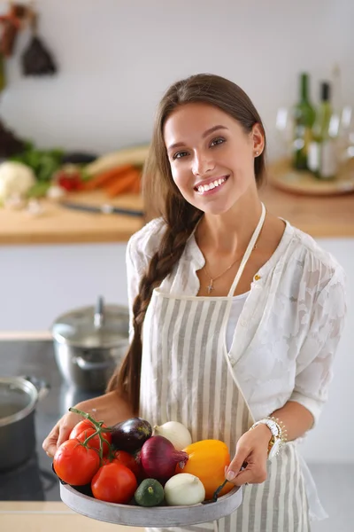 Smiling young woman holding vegetables standing in kitchen — Stock Photo, Image