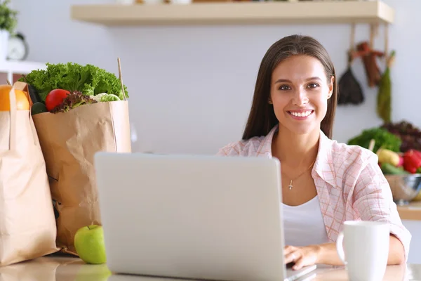 Smiling young woman with coffee cup and laptop in the kitchen at home — Stock Photo, Image