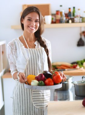Mutfakta elinde sebzelerle gülümseyen genç bir kadın.