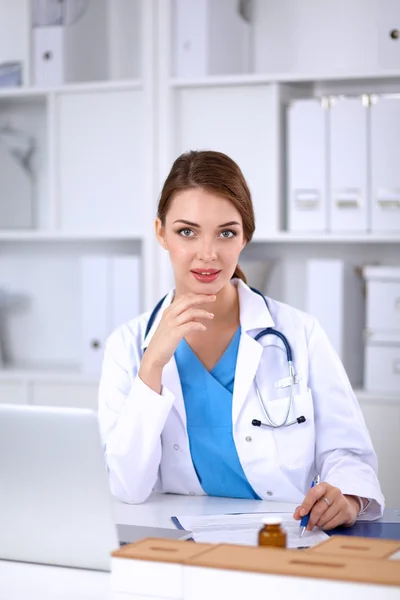 Beautiful young smiling female doctor sitting at the desk and writing. — Stock Photo, Image