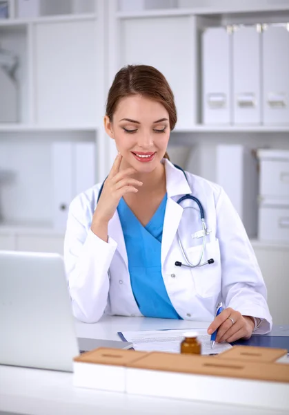 Schöne junge lächelnde Ärztin sitzt am Schreibtisch und schreibt. — Stockfoto